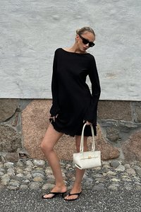 Vestido negro de manga larga con textura acanalada y dobladillo con volantes, combinado con chanclas negras y un pequeño bolso blanco. Textura suave y diseño minimalista.