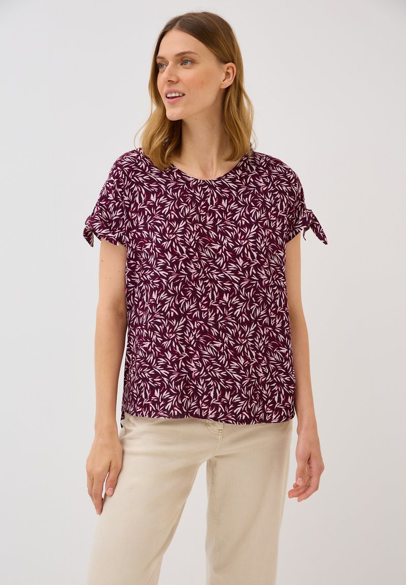Blouse bourgogne avec imprimé feuilles blanches, manches courtes avec accents de nœud, col rond et coupe décontractée, associée à un pantalon beige.