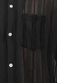 Černá košile na knoflíky vyrobená z texturované, průhledné tkaniny s vertikálními pruhy, s náprsní kapsou a krémovými knoflíky.