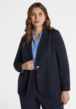 Blazer navy con botones dorados, diseño de un solo botón y solapas con muesca. Llevado sobre una blusa de color azul claro, con una textura suave y un corte ajustado.
