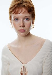 Cremer, gerippter Pullover mit V-Ausschnitt und Bindeschleife am Ausschnitt. Das Model hat kurzes, hellrotes Haar und trägt kleine Creolen.