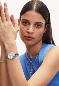 Silberner Armbanduhr mit runder Zifferblatt und texturiertem silbernen Band, getragen an einem nackten Arm. Das Modell trägt ein blaues, geripptes Tanktop.