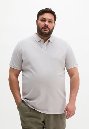 Mężczyzna z brodą, ubrany w jasnoszarą koszulkę polo i oliwkowe spodnie, stojący z jedną ręką w kieszeni na jednolitym tle.