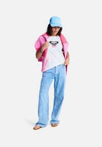 Hvit t-skjorte med svart Roxy-logo, rosa omslag, lyseblå caps, lyseblå vide jeans og brune sandaler på en modell mot en ensfarget bakgrunn.