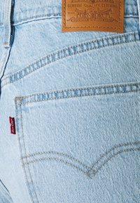 Vaqueros de mezclilla azul claro con un parche de cuero en la cinturilla, con un destacado detalle de costura en zigzag en el bolsillo trasero.