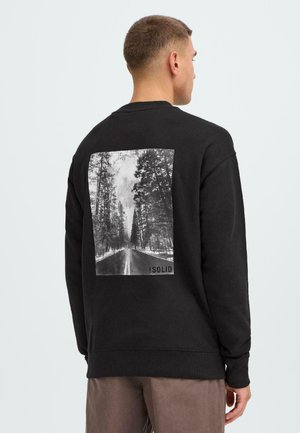 Man wearing black sweatshirt with black-and-white forest road photo and "!SOLID" text on the back, facing away, with brown pants.
