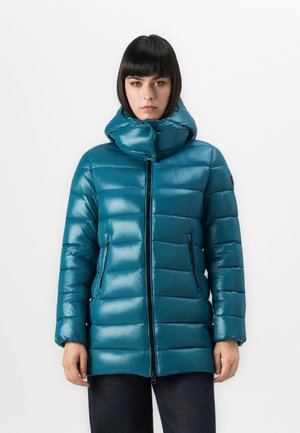 Vrouw in een glanzende teal pufferjack met hoge kraag, ritssluiting aan de voorkant en ritszakken aan de zijkanten, tegen een witte achtergrond.