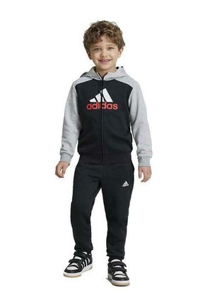Chándal adidas en negro y gris con sudadera con cremallera y pantalones a juego. Presenta el logo de adidas en rojo. Zapatillas negras con suelas blancas.