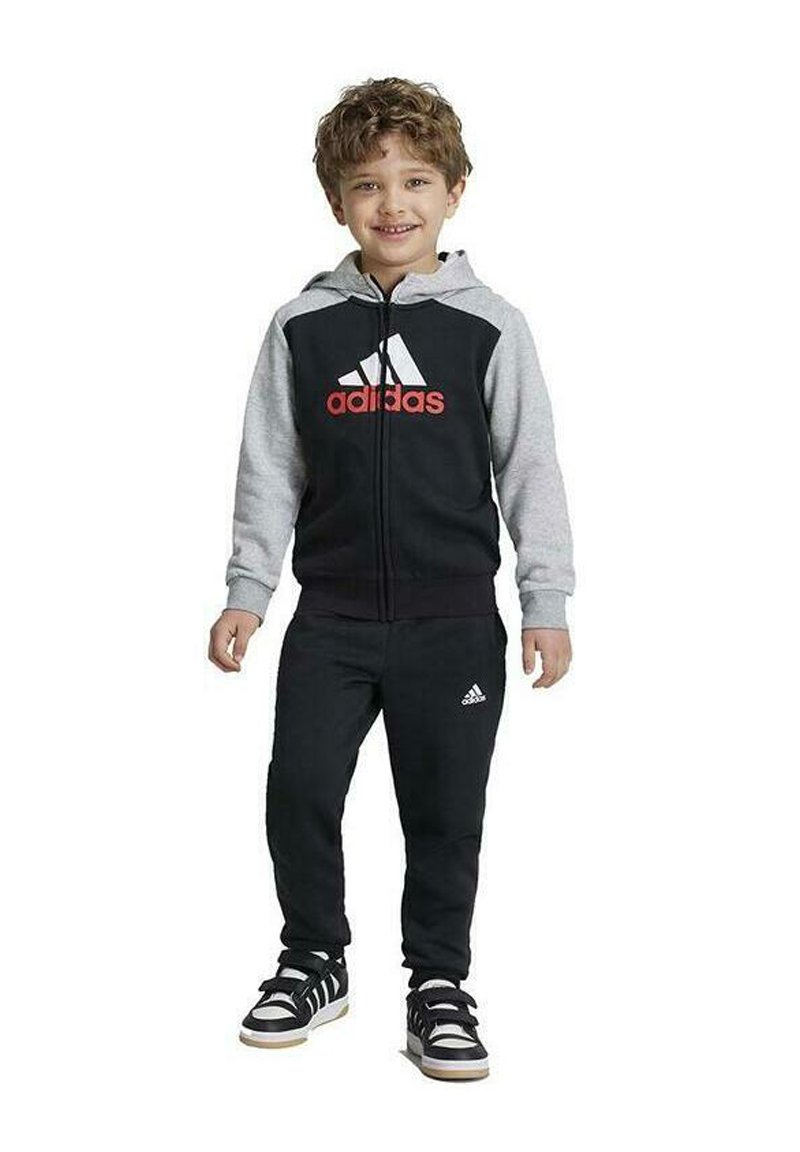 Tuta Adidas nera e grigia con felpa con zip e pantaloni coordinati. Presenta il logo Adidas in rosso. Sneakers nere con suole bianche.