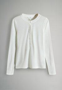 Valge pika varrukaga henley-särk, mille eesmine nööpide rida sisaldab viit nööpi, ripub lihtsa heledashall tausta ees.