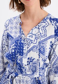 Vestido de estampado azul y blanco con un tejido suave, botones en la parte delantera, cintura con cordón y mangas largas que presentan diseños florales y de paisley.