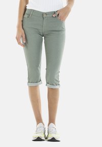 Pantalons capri vert clair en denim, avec des revers retroussés, une poche avant et une fermeture à bouton doré. Associés à des baskets blanches et grises.