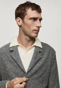 Gray wool blend coat with a notched lapel, layered over a cream textured knit polo. Person adjusts the coat's lapel, facing sideways.