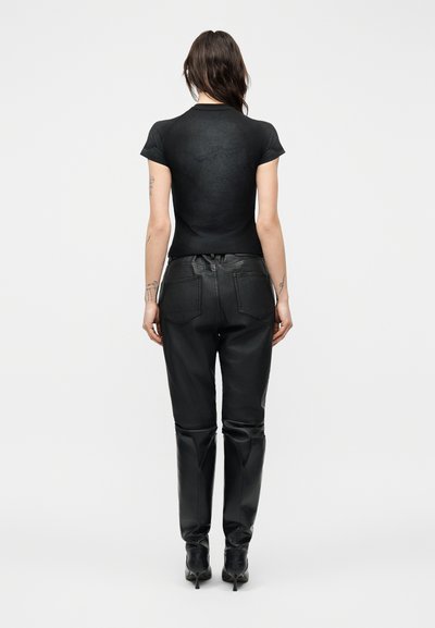 Γυναίκα που στέκεται γυρισμένη με την πλάτη προς την κάμερα, φορώντας μαύρο μπλουζάκι με κοντά μανίκια, μαύρο δερμάτινο παντελόνι και μαύρα μπότες με τακούνι, σε απλό φόντο.