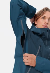 Teal softshell jack met grijze ritsen. Kenmerken zijn een hoge kraag, verstelbare manchetten en een ritszak op de borst. Glad, lichtgewicht materiaal.