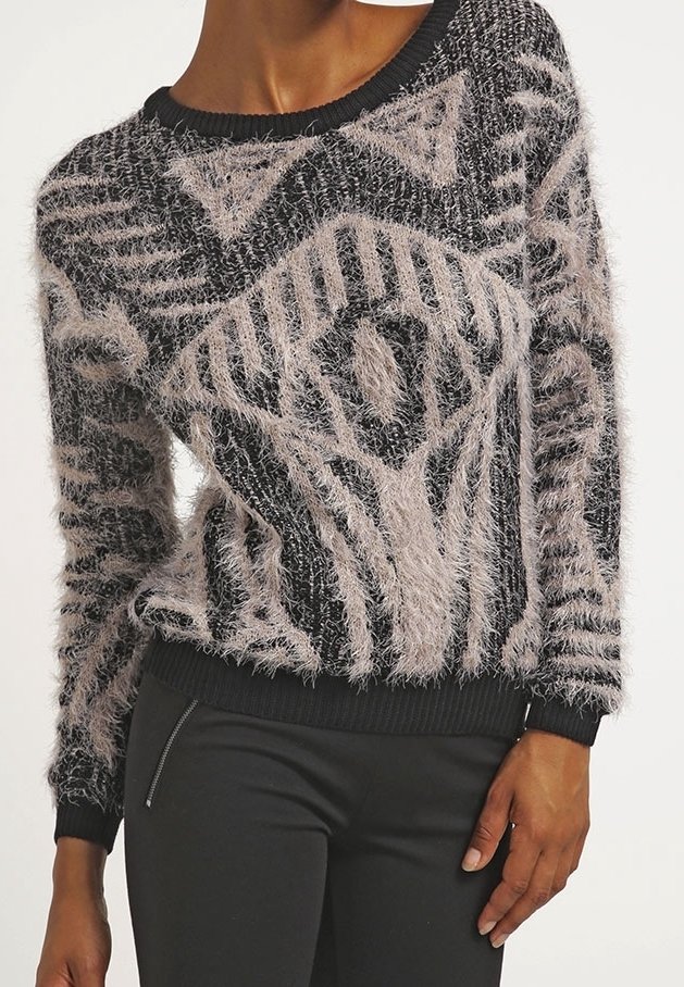 Pull doux noir et beige avec motif abstrait géométrique porté par une personne avec la main posée sur la hanche, assorti à un pantalon noir.