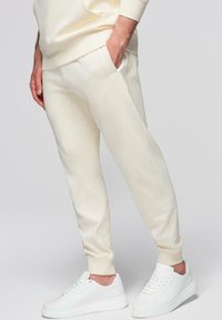 Homme portant un pantalon de jogging crème et des baskets blanches, debout avec une main dans la poche devant un fond uni.