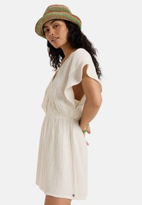 White, textured dress with flutter sleeves and a cinched waist, paired with a multicolored woven hat. Green bracelet accents the wrist.