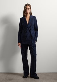Una donna è in piedi contro un muro bianco indossando un blazer doppiopetto blu navy e pantaloni a gamba larga coordinati, con mocassini neri.