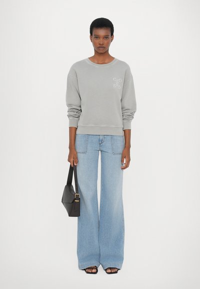 Felpa grigia con scollo rotondo e testo ricamato, abbinata a jeans a gamba larga di colore azzurro chiaro. Il modello tiene in mano una borsa nera.
