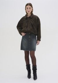 Brun läderjacka med krage, kombinerad med en blå denimkjol. Modellen bär svarta knähöga, rynkade klackstövlar och mörka strumpbyxor.