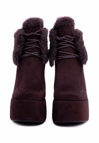 Stivali alla caviglia in suede marrone scuro con interno in finto pelliccia, punta rotonda, chiusura con lacci e suola spessa e piatta. La texture appare morbida e soffice.