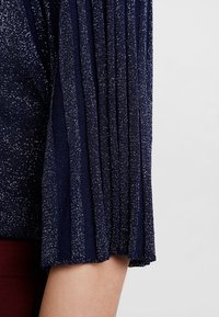 Marineblaues Oberteil mit glitzernder Textur und plissierten Ärmeln, das eine Kombination aus glattem und strukturiertem Stoff zeigt.