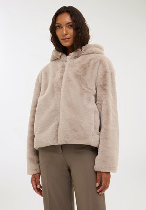 Giacca con cappuccio in pelliccia sintetica di un beige chiaro, con una forma squadrata, dalla consistenza morbida e con zip frontale. Abbinata a pantaloni grigi sartoriali.