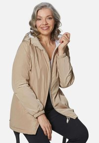 Beige ritsjack met capuchon, elastische manchetten en zijzakken, gemaakt van lichtgewicht materiaal, gedragen over een witte top.
