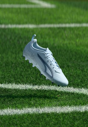 Chaussure de football blanche avec des détails bleus flottant au-dessus d'une pelouse verte marquée de lignes blanches.