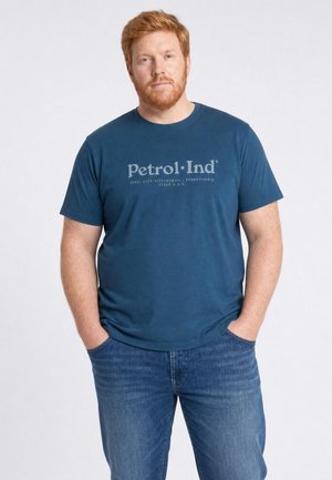 Hombre con pelo rojo y barba, vestido con una camiseta azul Petrol-Ind y vaqueros azules, de pie con las manos en los bolsillos frente a un fondo liso.