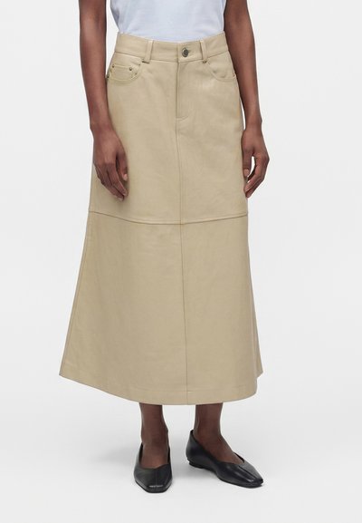 Jupe midi beige en ligne A avec poches devant et fermeture par boutons, portée avec des chaussures noires à petits talons et un haut blanc.