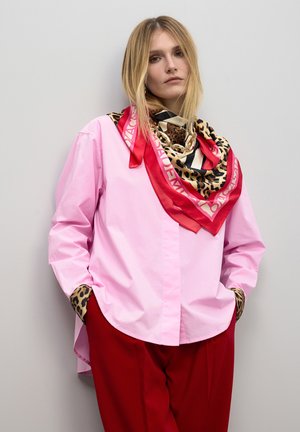 Camisa rosa de manga larga con puños de estampado de leopardo, combinada con una bufanda de múltiples patrones que presenta acentos en rojo, beige y negro. Pantalones rojos.