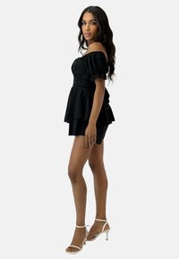 Top peplum nero con spalle scoperte, maniche corte, design a strati e pantaloni abbinati. La modella indossa tacchi argentati con cinturini.