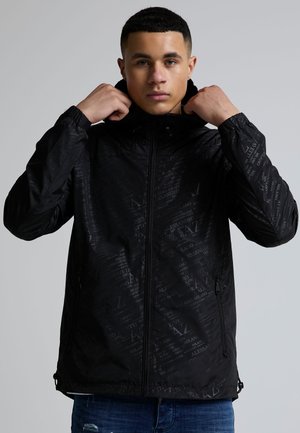 Ung mand justerer huen på en sort lynlåstrøje med subtile tekstmønstre, iført blå jeans mod en ensfarvet baggrund.