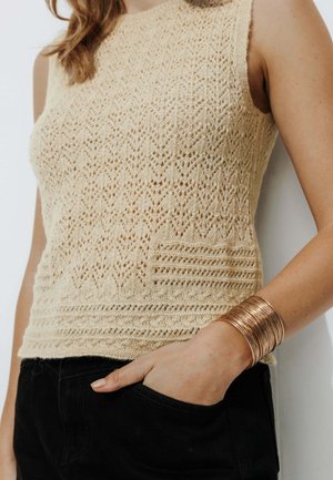 Un haut sans manches beige en tricot avec un motif en zigzag texturé et des accents côtelés à l'ourlet, associé à une main reposant dans une poche noire.