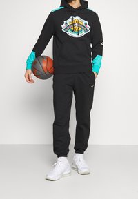 Schwarzer Hoodie mit türkisfarbenen Ärmeln und einem grafischen Logo, kombiniert mit schwarzen Jogginghosen und weißen Sportschuhen, während er einen Basketball hält.