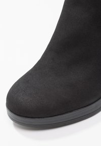 Bout de botte en daim noir avec une forme arrondie et une semelle épaisse en caoutchouc noir sur fond blanc.