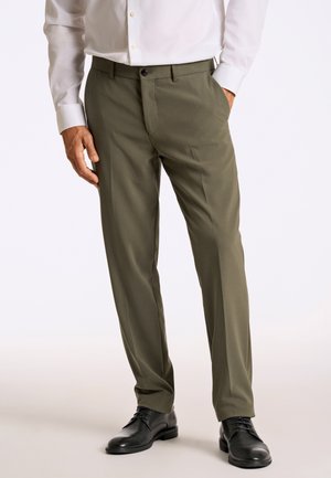 Uomo che indossa pantaloni eleganti verde oliva, camicia bianca infilata, scarpe eleganti nere, in piedi con una mano in tasca.