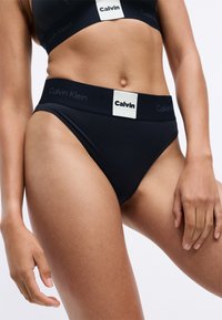 Svart Calvin Klein bikini med bred elastisk midjebånd som har et rektangulært logo-lapp. Stoffet ser glatt og kroppsnært ut.