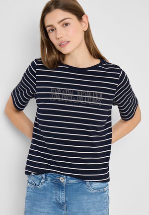 Femme aux cheveux bruns portant un T-shirt à rayures bleu marine et blanc avec le texte "BONJOUR" et un jean bleu clair, debout devant un fond uni.