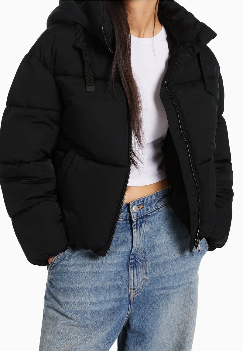 Černá péřová bunda s kapucí, vybavená elastickými šňůrkami, zipovým zapínáním a bočními kapsami, nošená přes bílý cropped top a modré džíny.