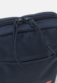 Sacca in tessuto blu navy con chiusura a zip, dotata di due tiranti in corda e una tasca frontale con cuciture in vista e un accento logo.