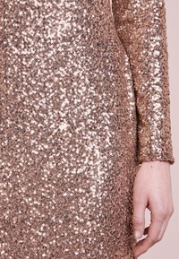Närbild av en hand bredvid ett långärmat klänningsstoff i roséguld med reflekterande, tätt packade paljetter.