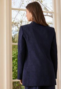 Navy blue jacket with a textured leaf pattern, structured shoulders, a tailored fit, and long sleeves, viewed from the back.