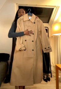 Femme tenant un trench-coat beige sur un cintre dans une chambre avec une lampe, un lit et des rideaux noirs en arrière-plan.