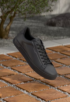 Basket en cuir noir avec un orteil arrondi, surface texturée et coutures ton sur ton. Elle présente des lacets plats noirs et un logo sur le côté.