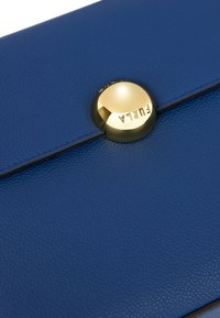 Oggetto in pelle blu con texture, dotato di un tasto rotondo e lucido in oro sul quale è impresso la parola "FURLA" su una linea di piega.
