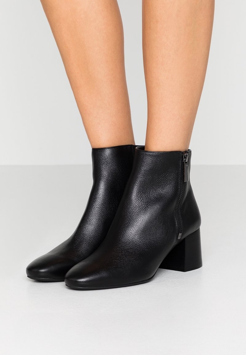Bottes en cuir noir à la cheville avec fermetures éclair latérales et talons blocs portées sur des jambes nues contre un fond uni.