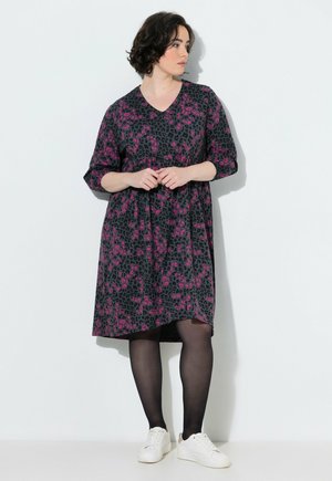 Donna con capelli corti e scuri che indossa un vestito fantasia nero e rosa al ginocchio, calze nere e scarpe da ginnastica bianche, in piedi contro un muro bianco.